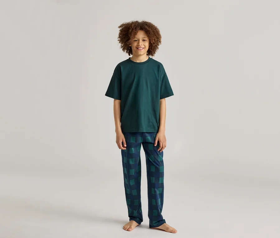 Ein lächelnder Junge mit lockigem Haar trägt einen dunkelgrünen Pyjama mit karierter Hose.