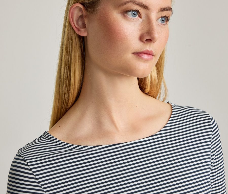 Nahaufnahme einer Frau mit blonden Haaren und blauen Augen, die ein dunkelblau-weiß gestreiftes Langarmshirt mit U-Boot-Ausschnitt trägt.