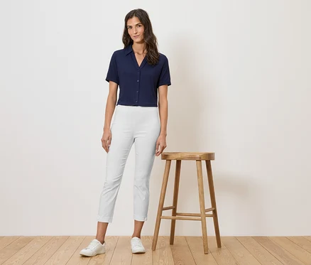 Frau in marineblauem Hemd und weißen Hosen, stehend neben einem Holzhocker.