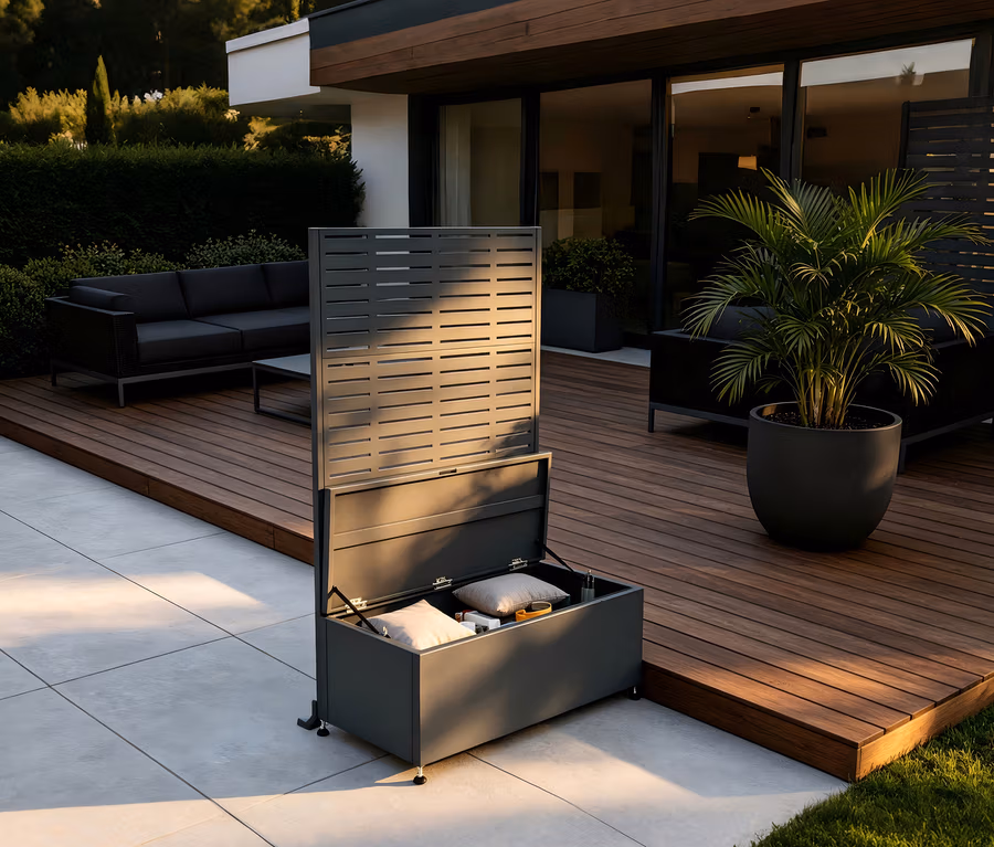 Dunkelgraue Outdoor-Aufbewahrungsbox mit offenem Deckel und integriertem Lamellen-Sichtschutz auf einer Terrasse. Kissen sind in der Box. Dahinter befindet sich eine Holzterrasse mit Sofa und Topfpflanze.