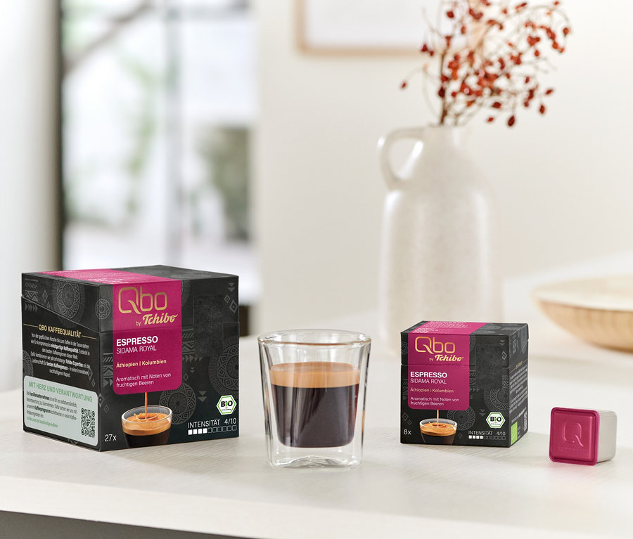 Zwei schwarz-pinke Qbo Espresso SIDAMA ROYAL Kapselpackungen (27 und 8 Kapseln), ein Glas Espresso und eine einzelne pink-weiße Kapsel auf einem weißen Tisch.