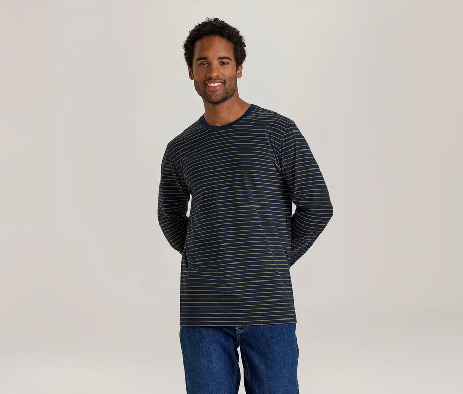 Ein lächelnder Mann mit dunklen Locken steht vor einem hellen Hintergrund und trägt ein dunkelblaues Langarmshirt mit dünnen beigen Streifen und Jeans.