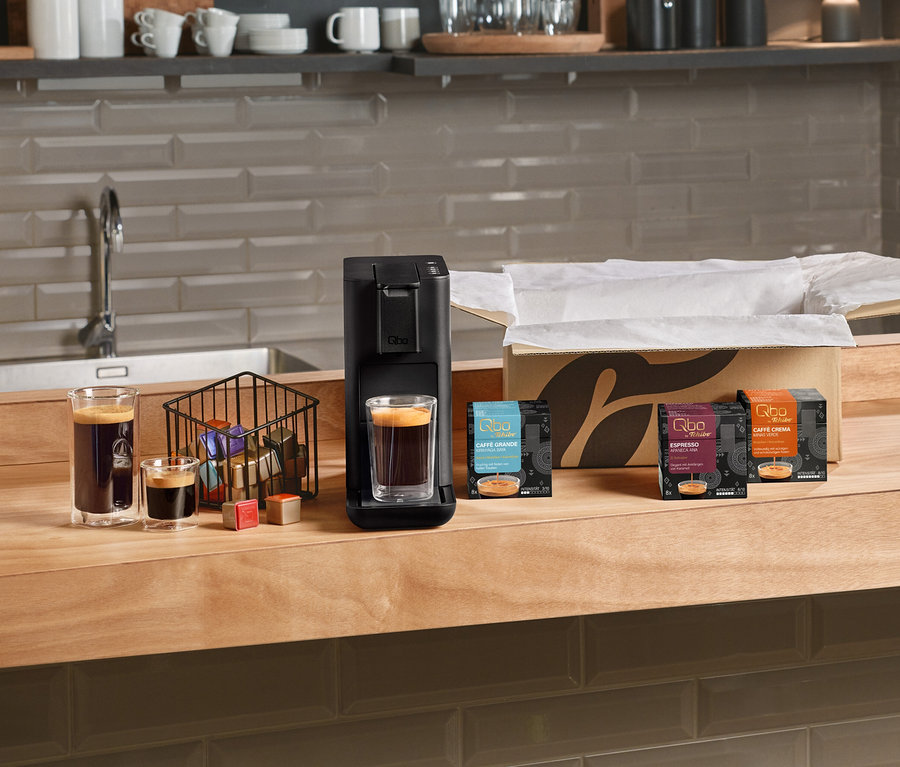 Eine schwarze Qbo Iconic Dark Chrome Kaffeemaschine mit einem Glas Espresso, daneben ein Korb mit Kapseln und drei Packungen Qbo Caffè Grande Kirinyaga Baya, Qbo Espresso Apaneca Ana und Qbo Caffè Crema auf einer Holztheke. Zwei weitere Gläser Kaffee stehen links.