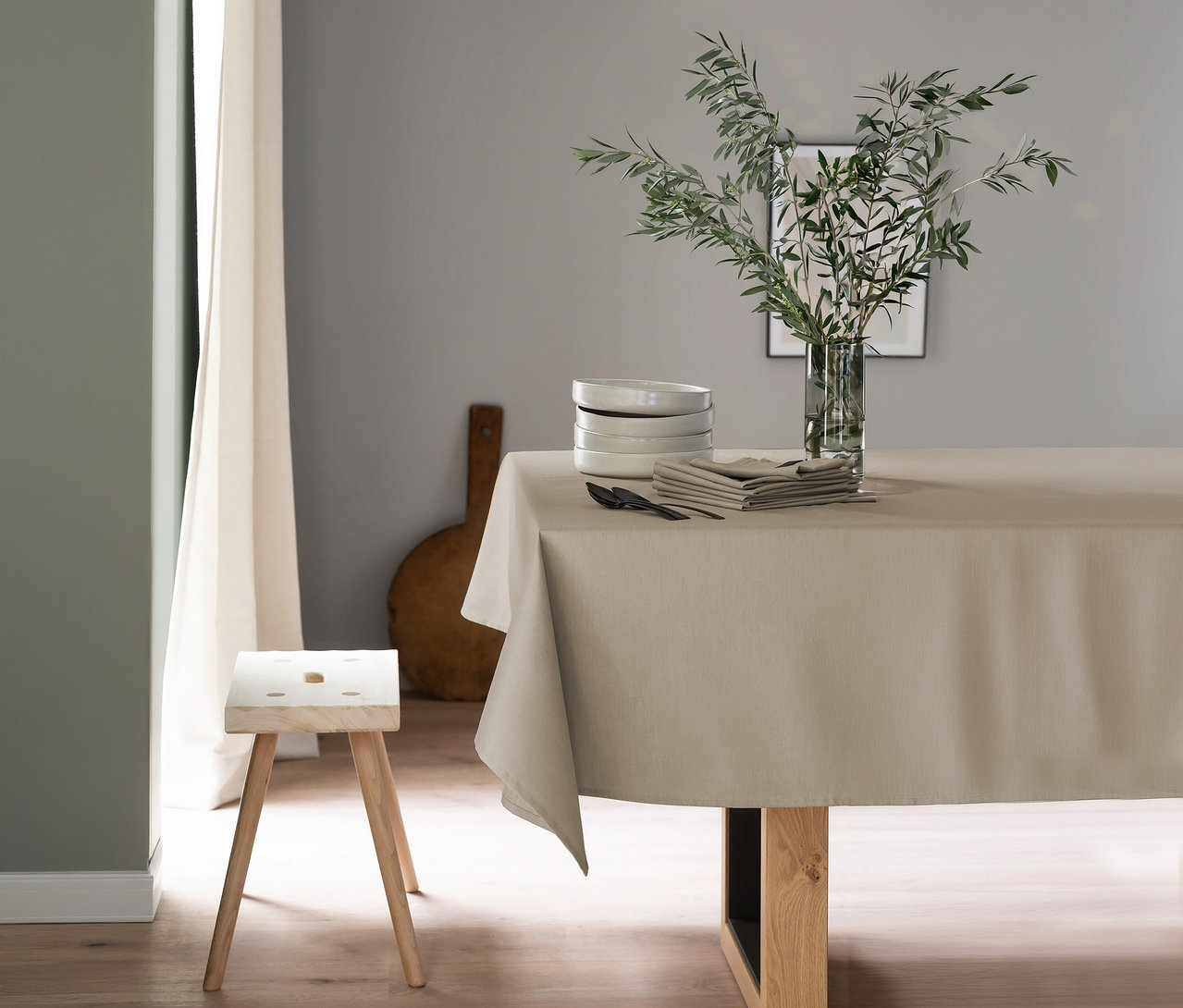 Ein Esstisch ist mit einer sandfarbenen SCHÖNER WOHNEN-Kollektion Tischdecke in Übergröße bedeckt. Darauf stehen weiße Schüsseln, sandfarbene Servietten, schwarzes Besteck und eine Glasvase mit grünen Olivenzweigen. Links daneben steht ein heller Holzschemel.