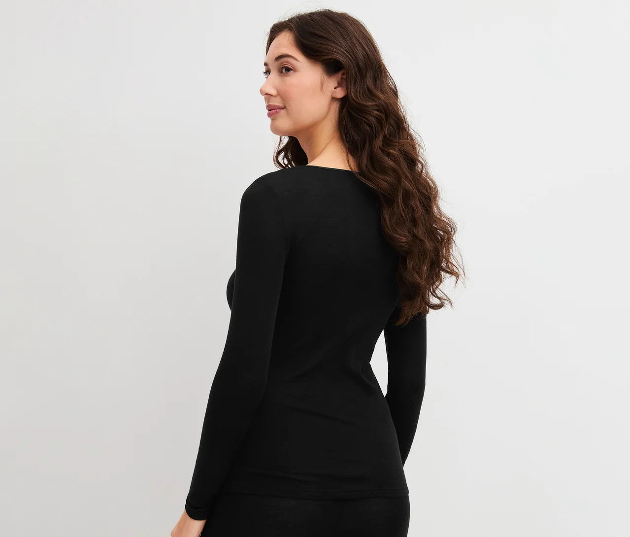 Frau mit langen, gewellten braunen Haaren trägt ein schwarzes CALIDA True Confidence Langarm-Shirt, von hinten gesehen.