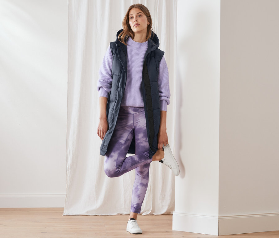 Frau posiert in langer Steppweste mit Kapuze, lila Pullover und Leggings mit lila Muster.