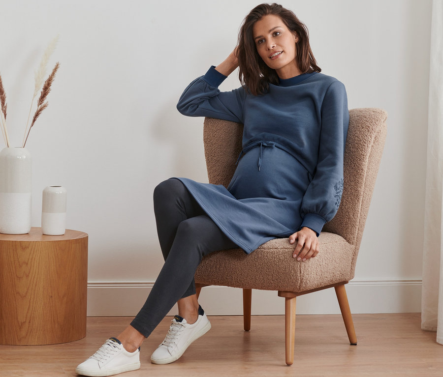Schwangere Frau sitzt im Sessel und trägt Still-/Umstands-Sweatkleid, Leggings und Sneakers.