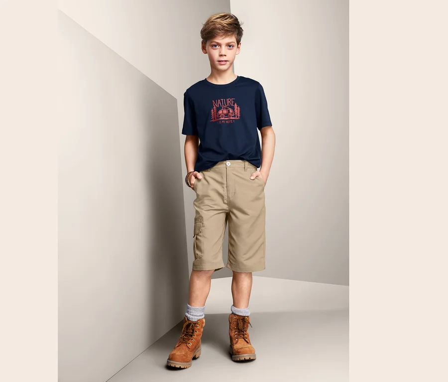 Ein Junge in einem bedruckten T-Shirt und beigen 2-in-1-Funktionsshorts steht in einer Ecke des Raumes.