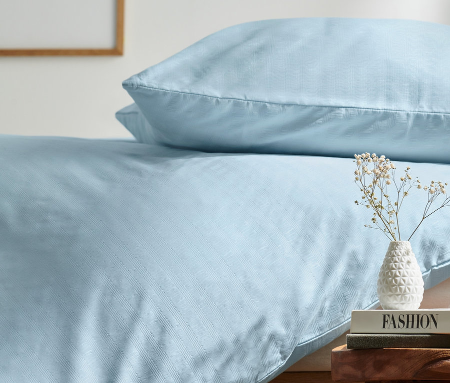 Hellblaue Seersucker-Bettwäsche auf einem Bett neben einem Holzhocker mit Büchern und einer Vase.
