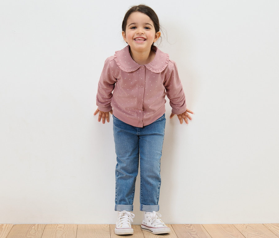 Lächelndes Mädchen steht vor einer weißen Wand und trägt Jeans und eine Kinder-Sweatjacke mit Volant.