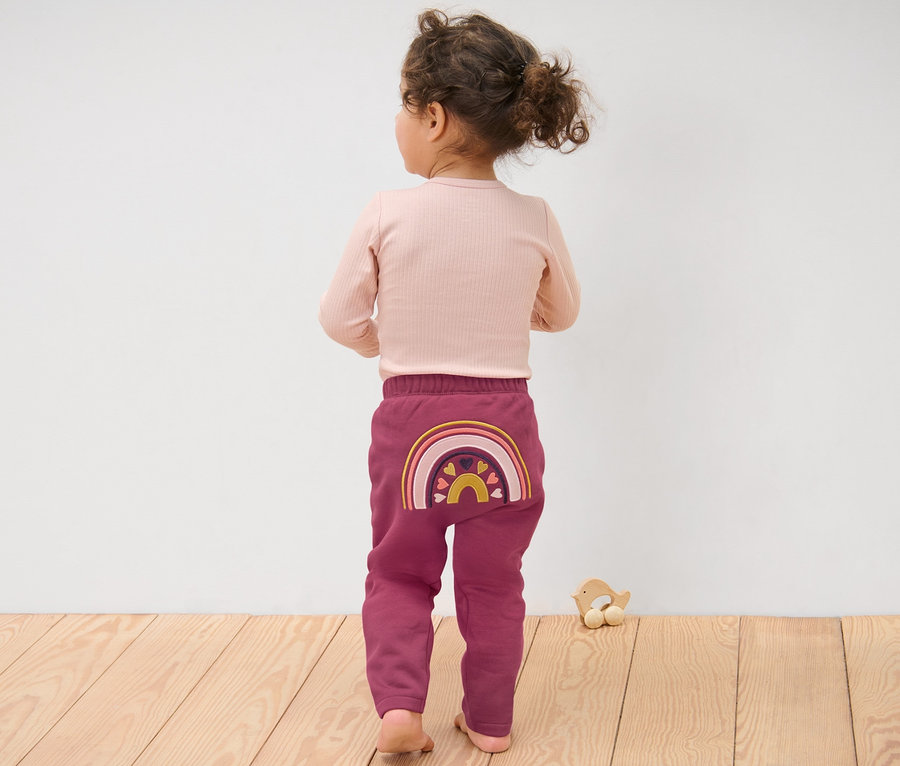 Mädchen in Baby-Sweathose, Regenbogen steht mit dem Rücken zur Kamera.