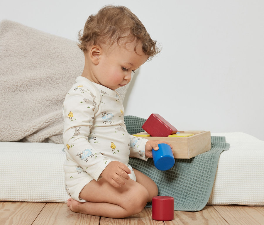 Ein lockiges Kleinkind sitzt auf dem Boden und spielt mit bunten Bauklötzen. Es trägt blaue 3 Baby-Bodys mit Mitwachsfunktion.