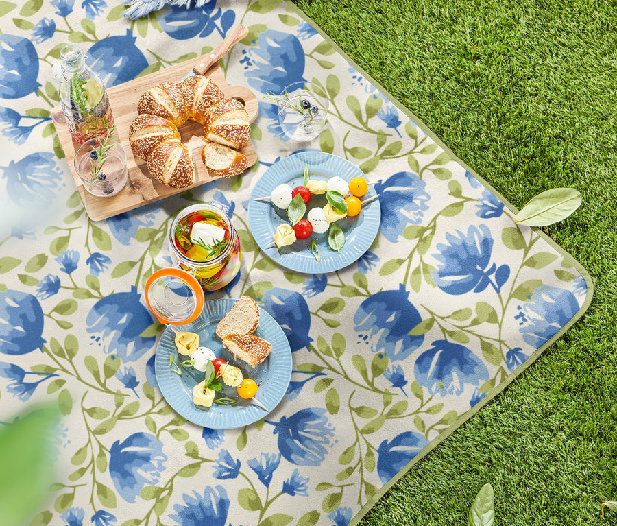 Auf dem Rasen liegt eine kleine Picknickdecke mit Blumenmuster. Darauf befinden sich Teller mit Essen, Gläser mit Getränken und ein Holzbrett mit Gebäck.