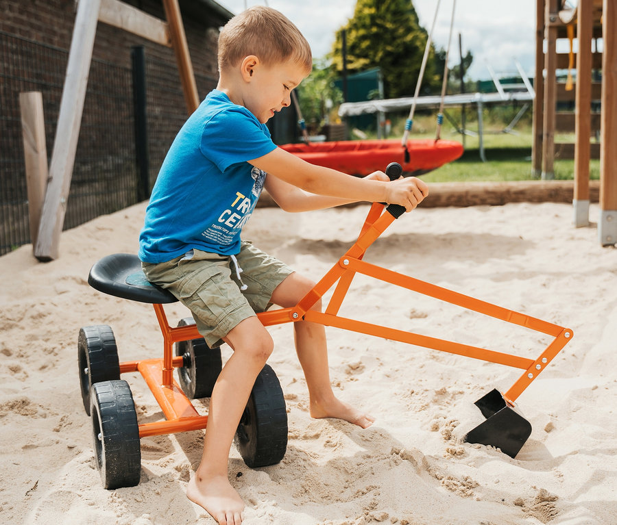 Ein Junge sitzt auf einem orangefarbenen small foot Bagger mit Rädern im Sand.