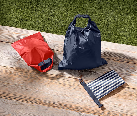 Ein roter, ein dunkelblauer und ein blau-weiss gestreifter wasserdichter Packsack liegen auf einer Holzfläche neben grünem Gras.