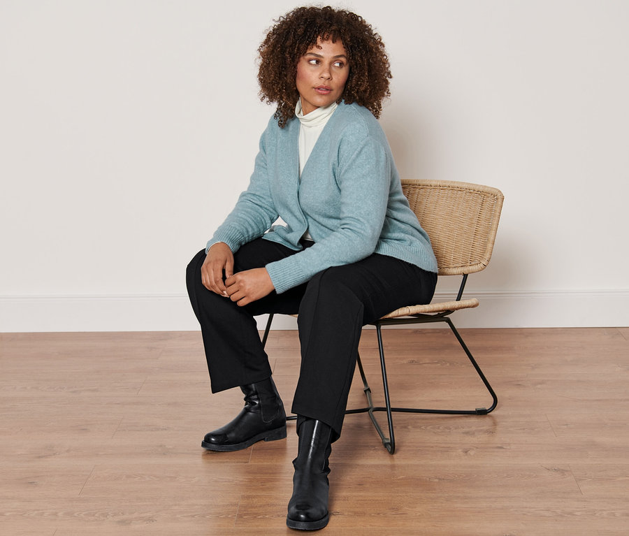 Frau sitzt auf einem Stuhl und trägt eine Strickjacke mit Wolle, ein cremefarbenes Rollkragenshirt, eine schwarze Jerseyhose mit Biese und Lederboots.