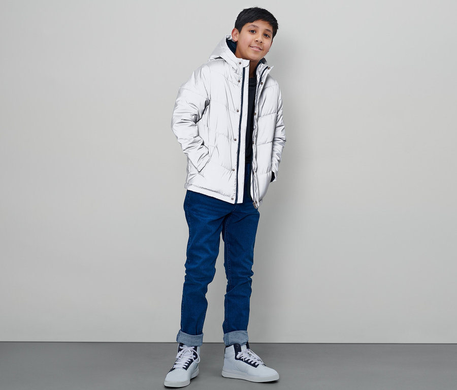 Ein Junge in einer reflektierenden Kinder-Steppjacke steht vor einer weißen Wand.