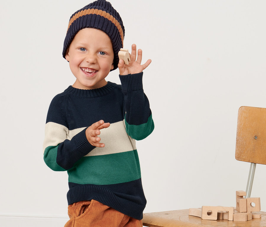 Ein Junge mit Strickpullover und Mütze hält ein Holzspielzeug.