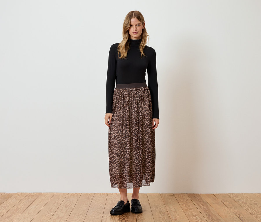 Frau steht in schwarzem Rollkragenoberteil, braunem Faltenrock mit Tiermuster und schwarzen Schuhen.