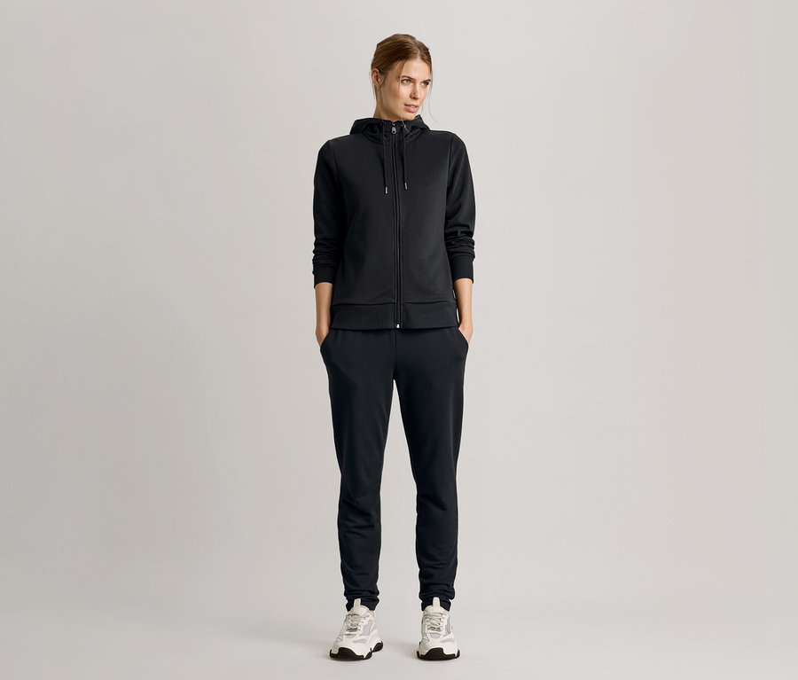 Frau steht mit Händen in den Taschen, trägt einen schwarzen Trainingsanzug mit Kapuzenjacke und Turnschuhe.