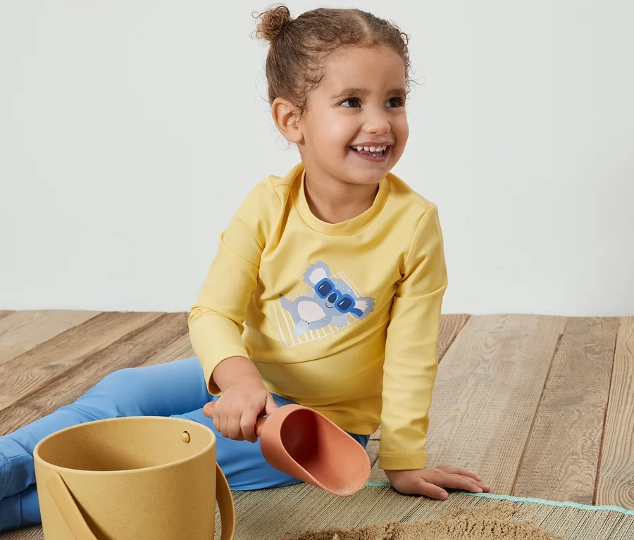 Lächelndes Mädchen sitzt auf dem Boden und spielt mit Sand in gelbem Kinder-Shirt mit UV-Schutzfaktor 80, Koala.