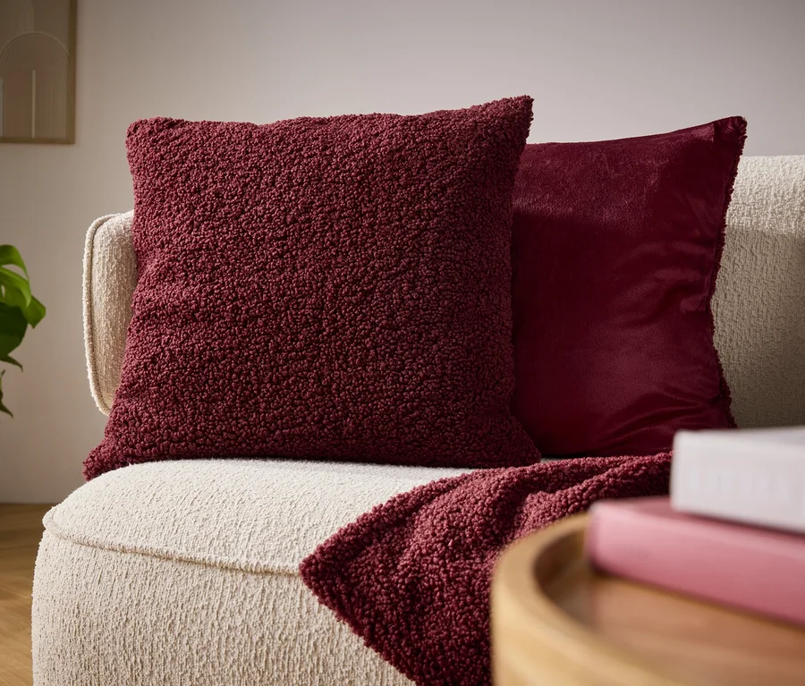 Detail eines beigen Sofas mit einem bordeauxroten Plüschkissen, einem glänzenden Kissen und einer drapierten Decke.
