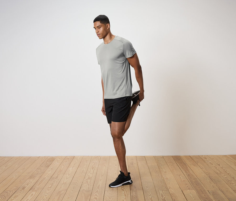 Mann in grauem T-Shirt und schwarzen Shorts dehnt sein Bein auf einem Holzboden.