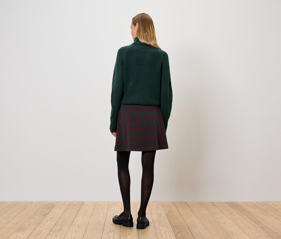 Eine Frau steht mit dem Rücken zur Kamera und trägt einen grünen Pullover, einen karierten Rock und eine schwarze Strumpfhose.