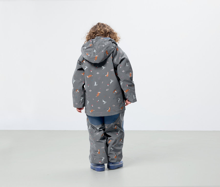Junge mit lockigen Haaren trägt eine graue Jacke und Hose mit Fuchs- und Vogelmuster, mit dem Rücken zur Kamera.