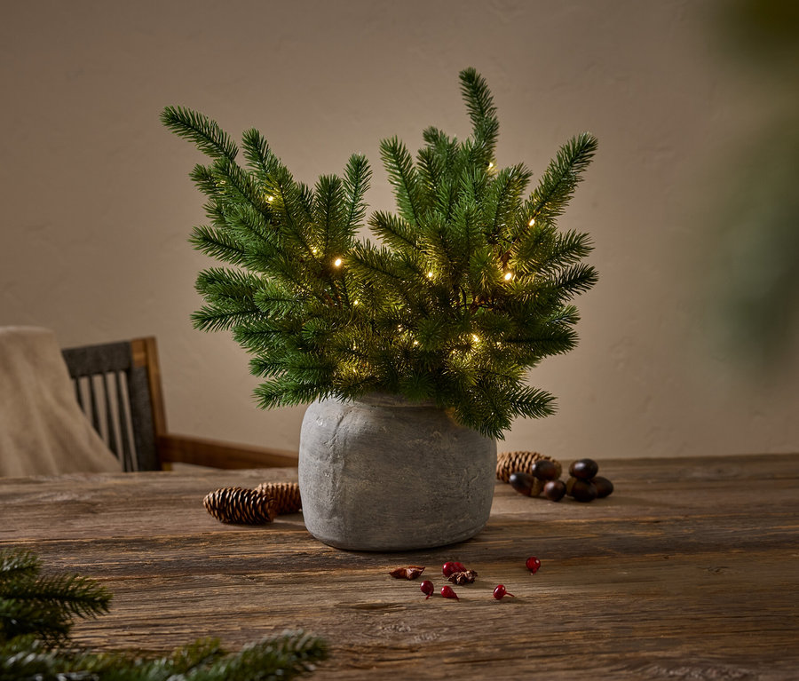 Grüner Nadelbaum mit kleinen Lichtern in einer grauen Vase auf einem Holztisch.
