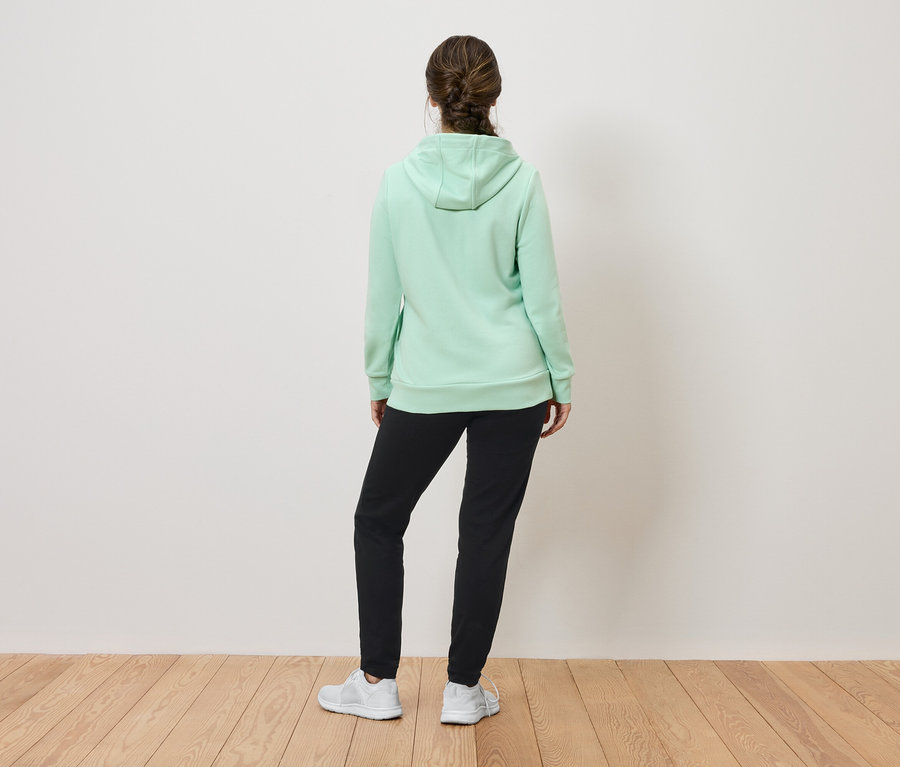 Frau mit dem Rücken zur Kamera trägt einen hellgrünen Hoodie, eine schwarze Jogginghose und weiße Turnschuhe.