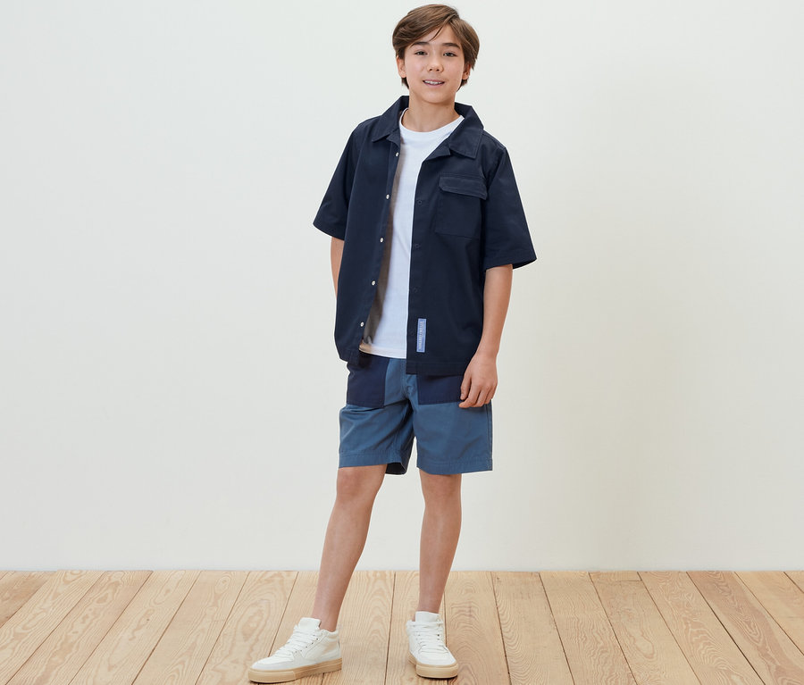 Ein Junge steht und trägt ein Kinder-Overshirt, Shorts und Turnschuhe.