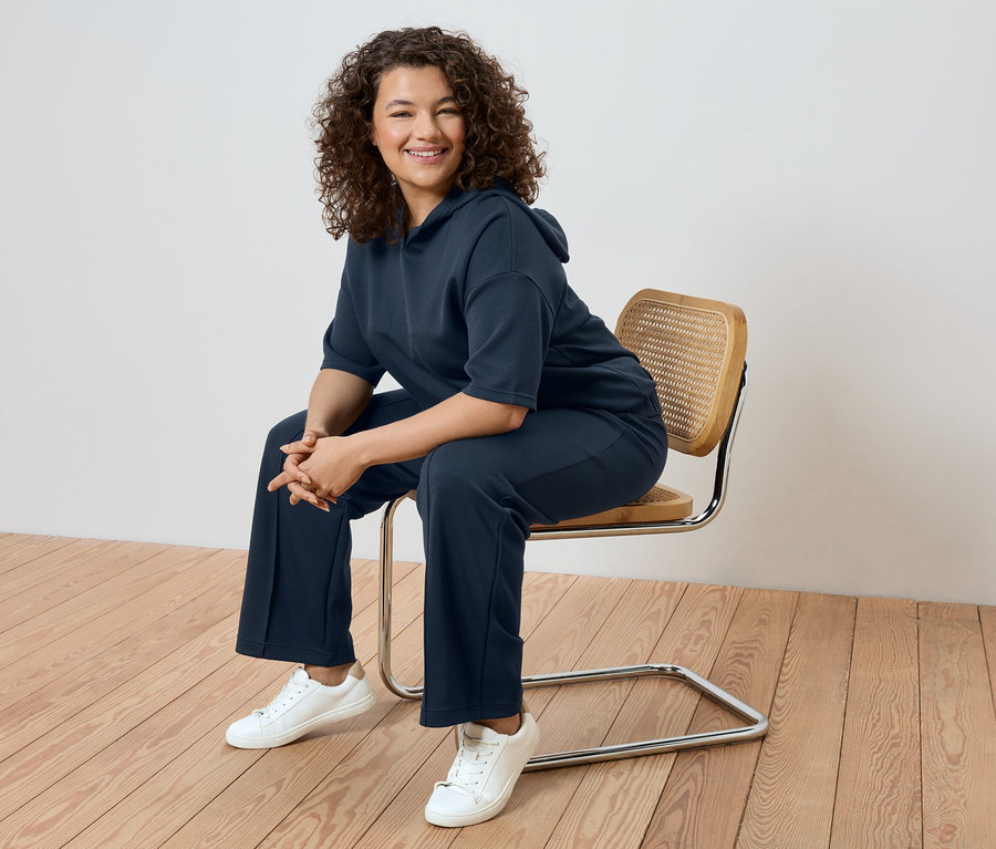 Frau mit lockigem Haar sitzt auf einem Stuhl und trägt einen marineblauen Trainingsanzug und weiße Turnschuhe.