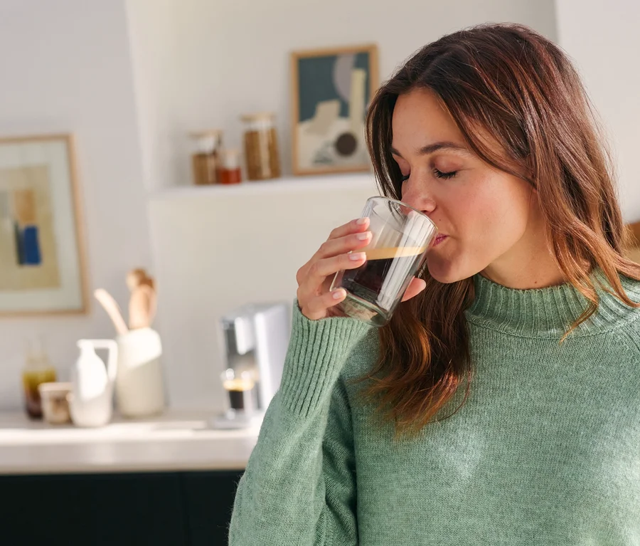 Eine Frau mit geschlossenen Augen trinkt Kaffee aus einem klaren Glas, trägt einen grünen Pullover, mit einer Kaffeemaschine auf einer Küchenzeile im Hintergrund.
