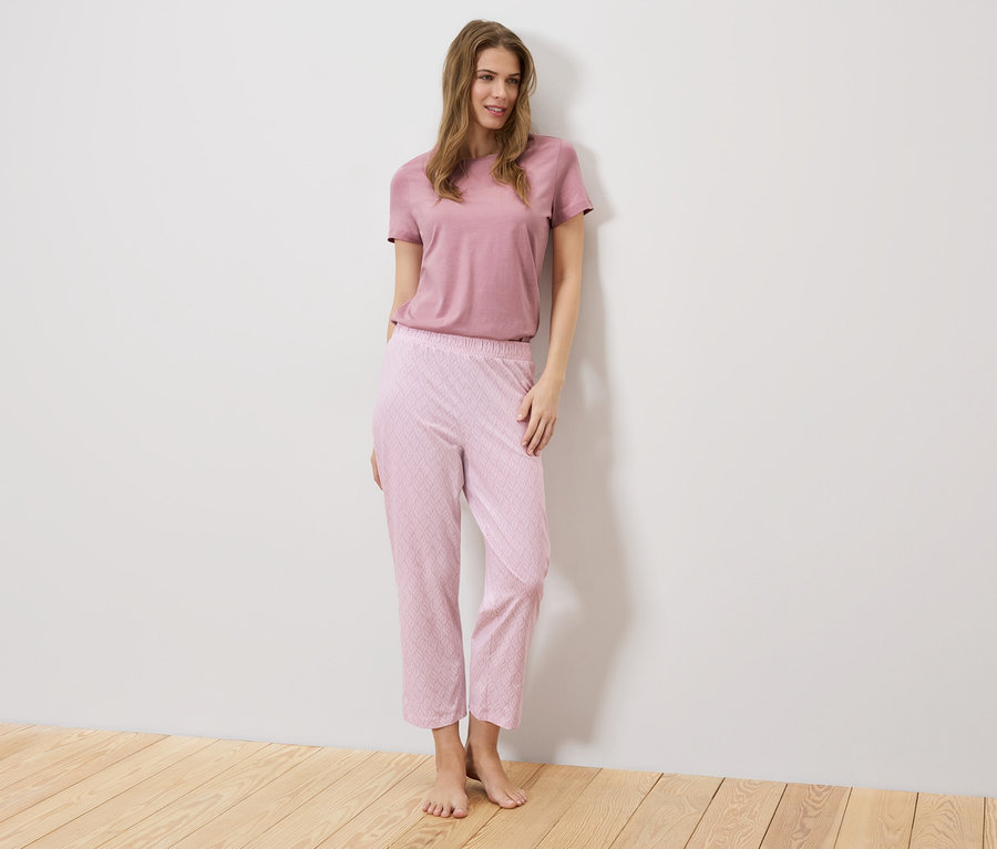 Eine Frau steht vor einer weißen Wand, sie trägt ein rosa T-Shirt und eine rosafarbene Hose mit Muster.