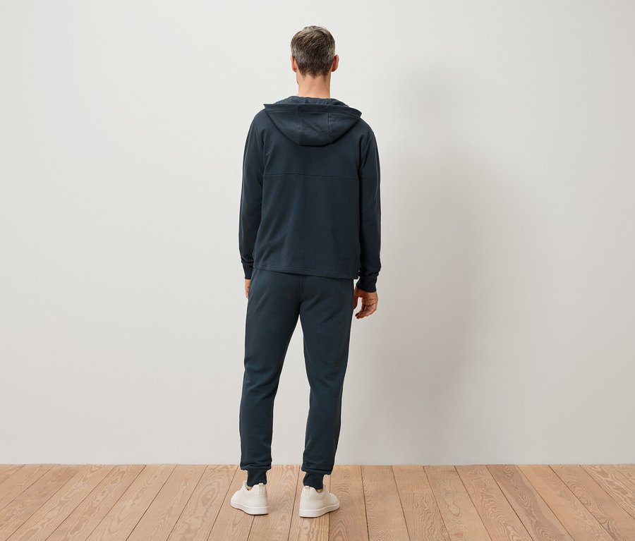 Rückansicht eines Mannes mit grauen Haaren, der einen dunkelblauen Hoodie und eine Jogginghose sowie weiße Turnschuhe trägt.