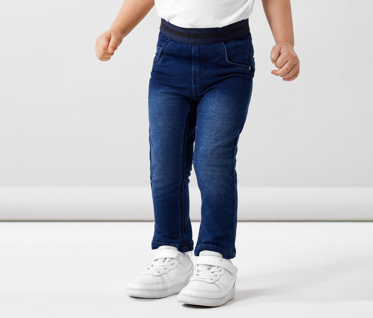 Kind trägt NAME IT Kinder-Sweat-Jeans und weiße Schuhe vor weißem Hintergrund.