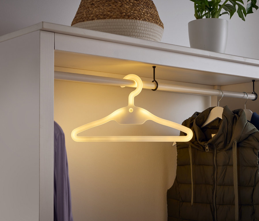 In einem weißen Kleiderschrank hängt ein leuchtender Kleiderbügel an einer Stange, neben einer Weste und einem Hemd.