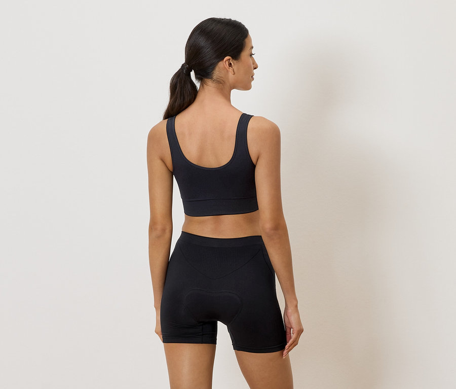 Frau von hinten in schwarzem Sport-BH und Radlerhose vor einem neutralen Hintergrund.