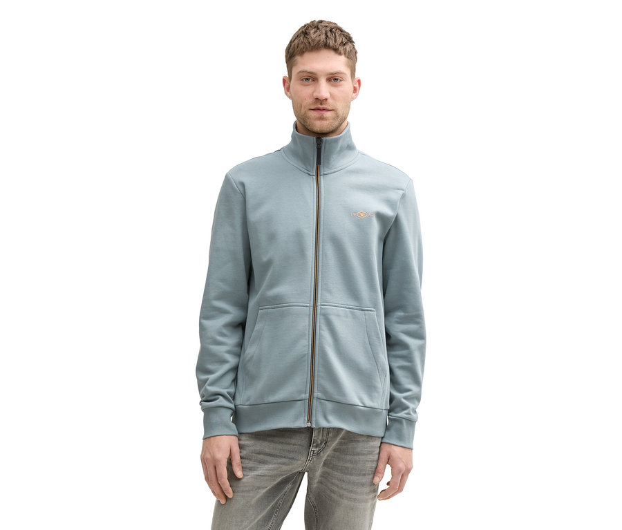 Mann trägt eine mintgraue Tom Tailor Sweatjacke mit Stehkragen und graue Jeans.