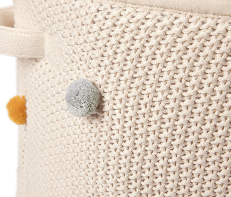 Detailansicht eines cremefarbenen, gestrickten Aufbewahrungskorbs mit Tragegriff und gelbem sowie grauem Bommel.