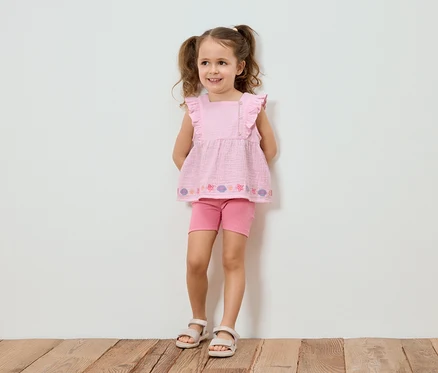 Mädchen mit Zöpfen trägt ein rosa Top mit Rüschen, Shorts und Sandalen, vor einer weißen Wand.