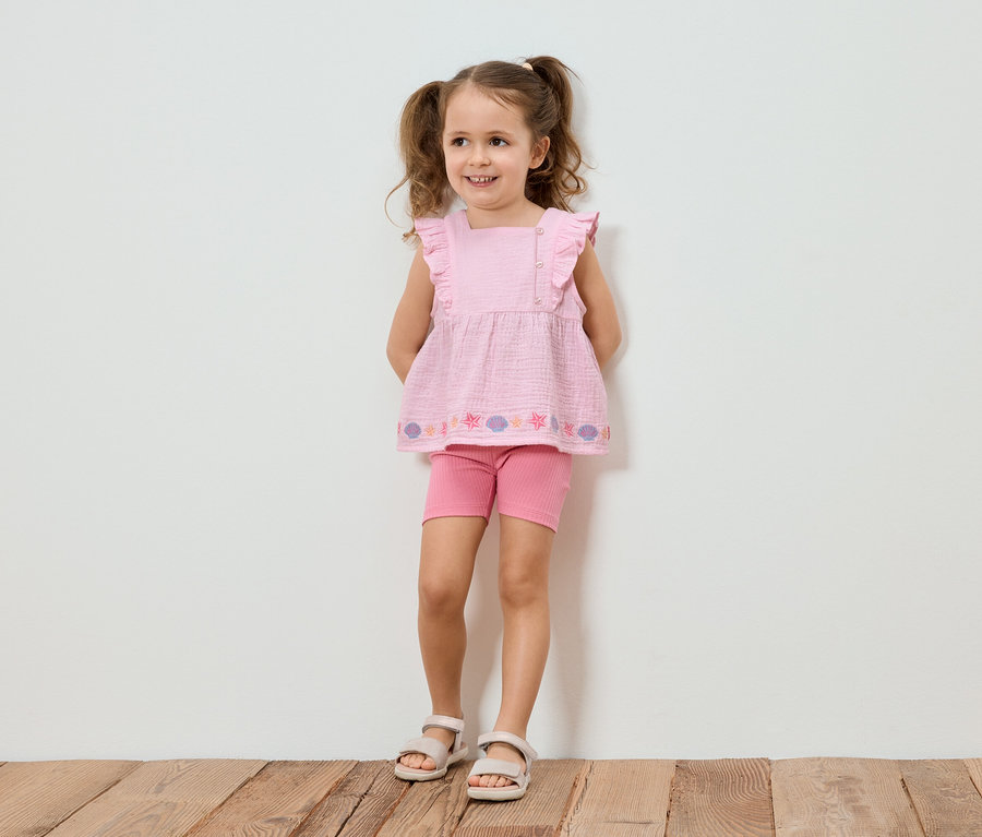 Mädchen mit Zöpfen trägt ein rosa Top mit Rüschen, Shorts und Sandalen, vor einer weißen Wand.