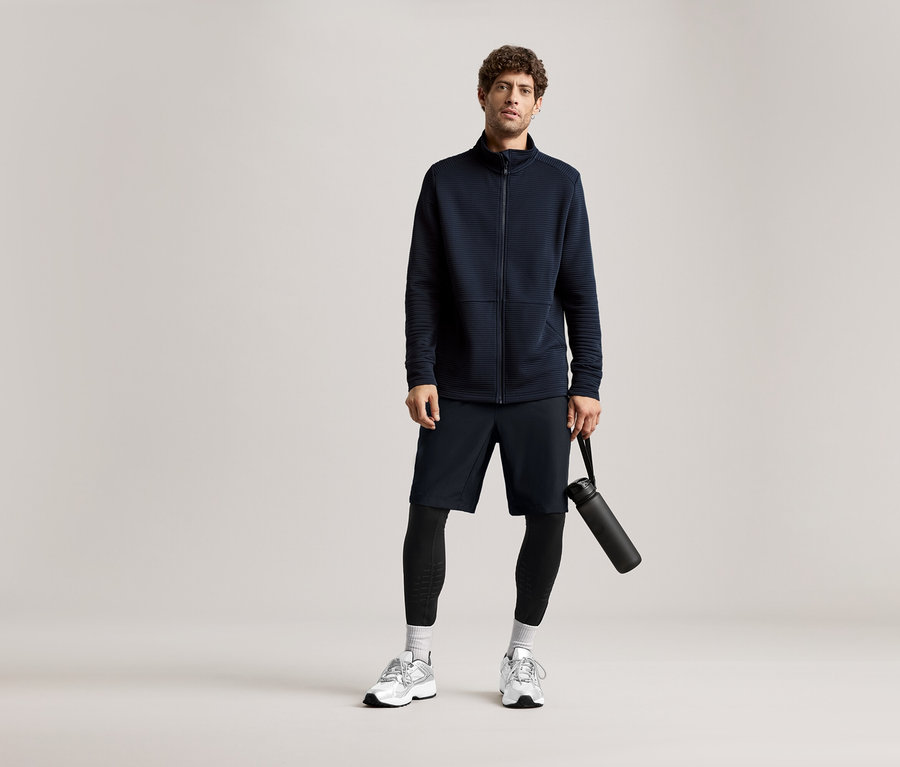 Mann in marineblauer Jacke, Shorts und Leggings, hält eine schwarze Wasserflasche.