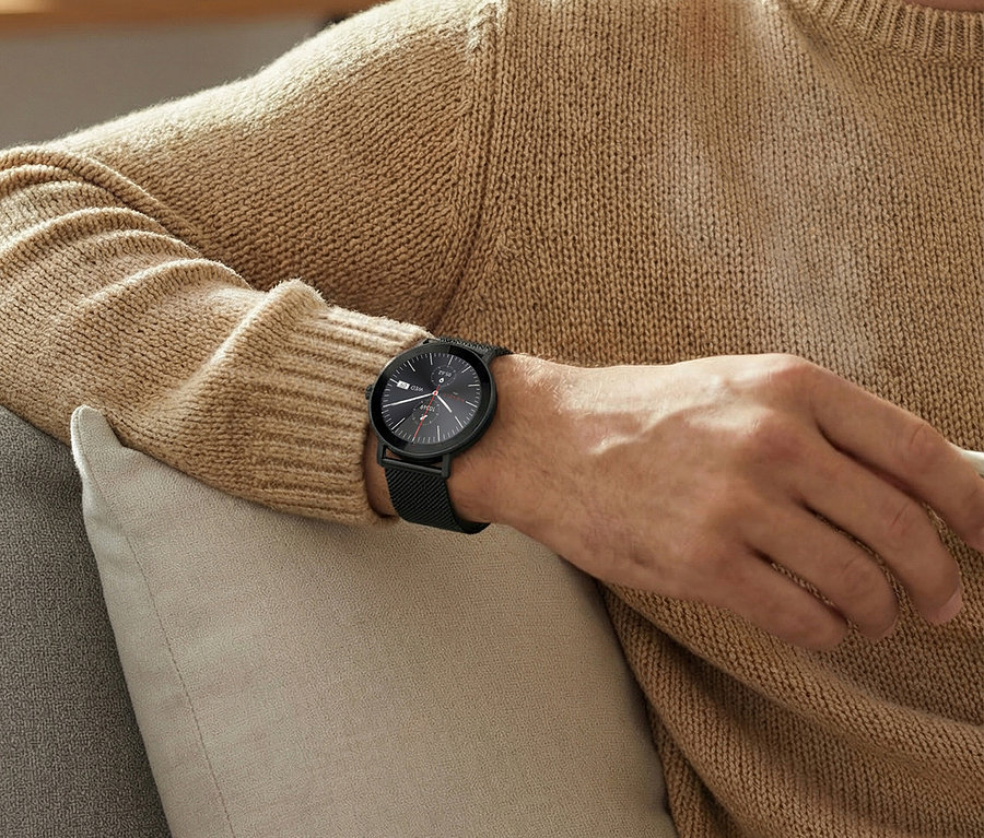 Mann mit beigem Pullover sitzt auf einem Sofa und trägt eine schwarze Armbanduhr am linken Handgelenk.