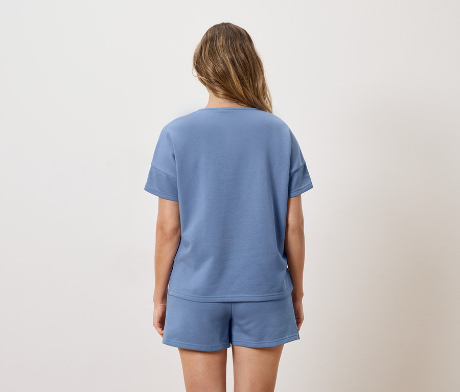 Rückansicht einer Frau mit langen, braunen Haaren, die ein blaues T-Shirt und Shorts trägt.