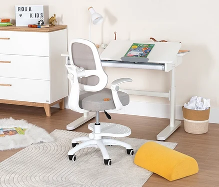 Ein Kinderzimmer zeigt einen Schreibtisch mit Stuhl, Lampe und Deko. Neben dem Schreibtisch steht eine Kommode und ein Papierkorb.