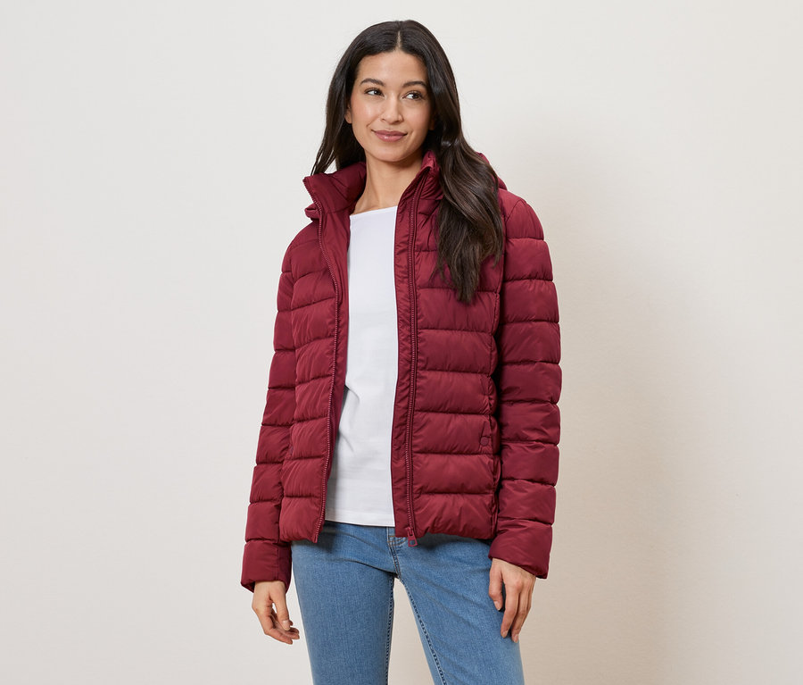 Frau mit langem, dunklem Haar trägt eine rote Steppjacke, ein weißes T-Shirt und Jeans vor einem hellen Hintergrund.