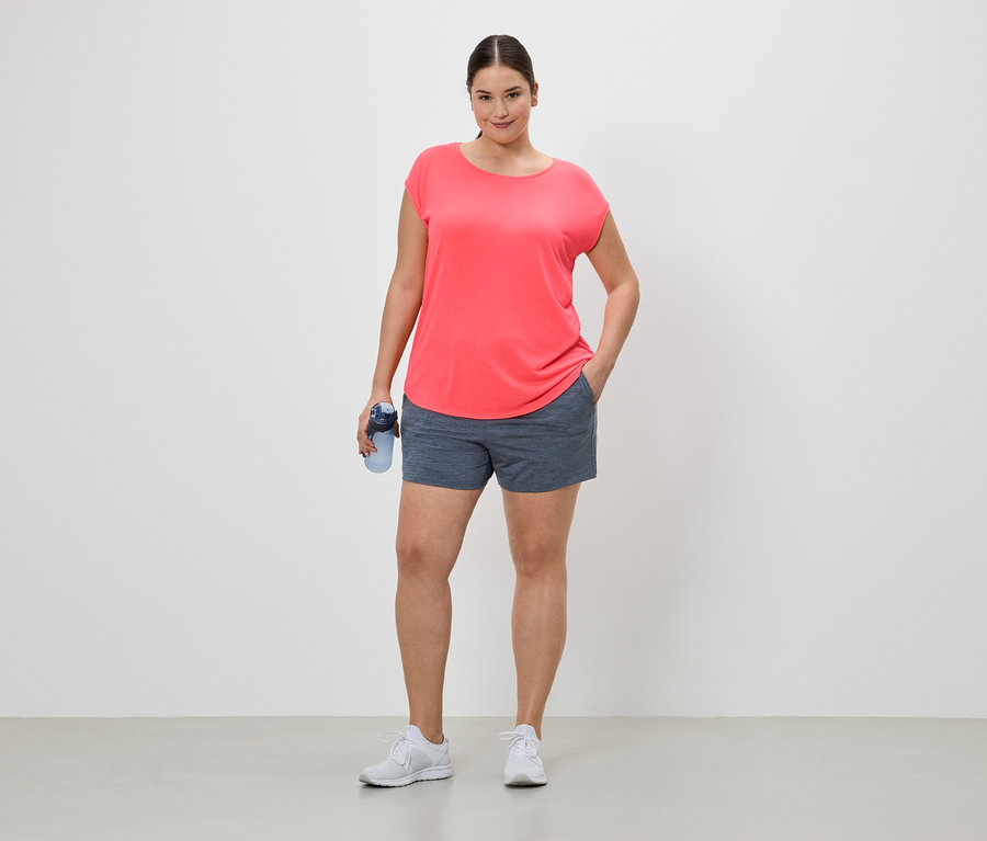 Frau in Sportkleidung mit Trinkflasche, stehend vor einem weißen Hintergrund. Sie trägt ein korallfarbenes T-Shirt, Shorts und weiße Turnschuhe.