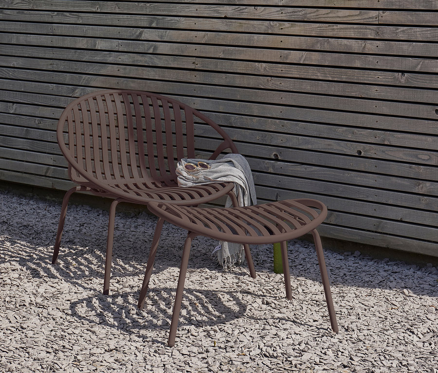 Brauner WOOOD Lounge Sessel mit Hocker steht auf Kies. Auf dem Sessel liegen eine Decke und eine Sonnenbrille.